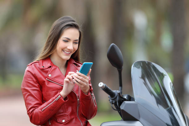 femme avec veste cuire rouge sur son telephone souriante a coté de son scooter